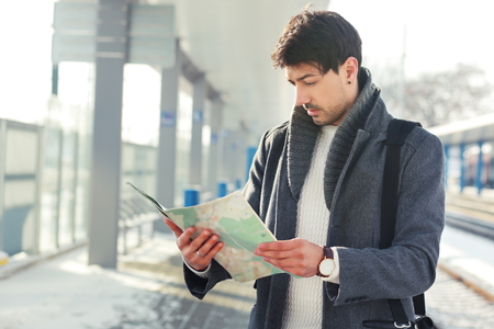 outdoor portrait of young handsome man standing at train station holding map in hands wearing warm coatの写真素材