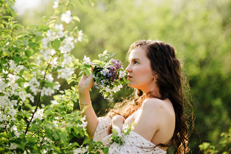 The young girl with a bouquet of wild flowers with naked shoulders in beams of the sun costs near a bush with white flowersの写真素材