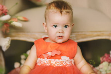the child in an orange dress with a big flower sits on a beige carpet and smilesの写真素材