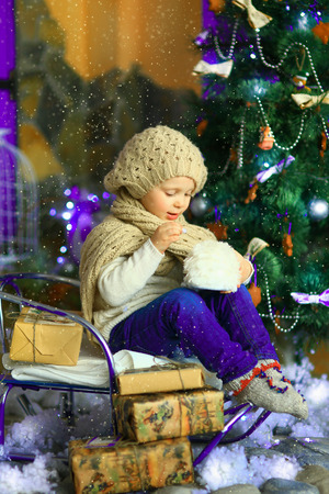 the girl in lilac jeans, in a knitted cap and with a knitted scarf sits on a sledge near a Christmas fir-tree with a toy and it is snowingの写真素材