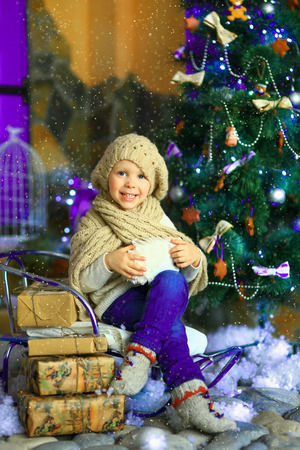 the girl in lilac jeans, in a knitted cap and with a knitted scarf sits on a sledge near a Christmas fir-tree with a toy and it is snowingの写真素材