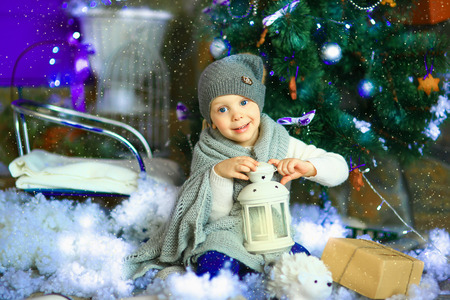 the girl in lilac jeans, in a knitted cap and with a knitted scarf sits on a sledge near a Christmas fir-tree with a toy and it is snowingの写真素材