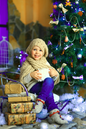 the girl in lilac jeans, in a knitted cap and with a knitted scarf sits on a sledge near a Christmas fir-tree with a toy and it is snowingの写真素材