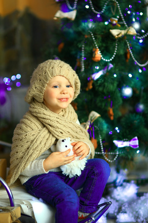 the girl in lilac jeans, in a knitted cap and with a knitted scarf sits on a sledge near a Christmas fir-tree with a toy and it is snowingの写真素材