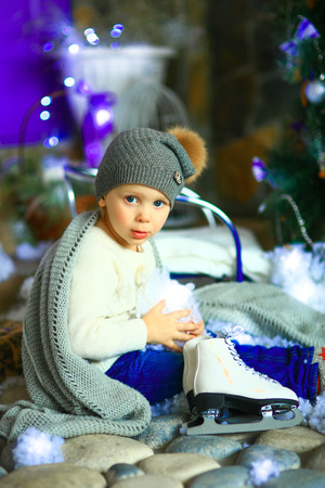 the girl in lilac jeans, in a knitted cap and with a knitted scarf sits on a sledge near a Christmas fir-tree with a toy and it is snowingの写真素材