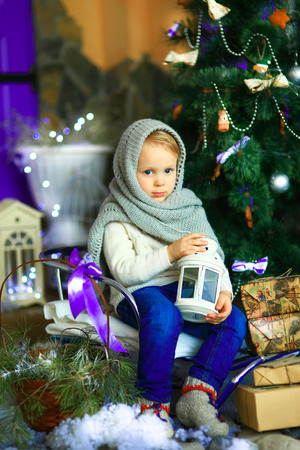 the girl in lilac jeans, in a knitted cap and with a knitted scarf sits on a sledge near a Christmas fir-tree with a toy and it is snowingの写真素材