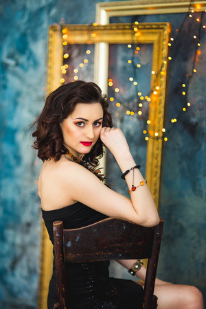 the girl with black hair in a black dress and in costume jewelry poses on a chairの写真素材
