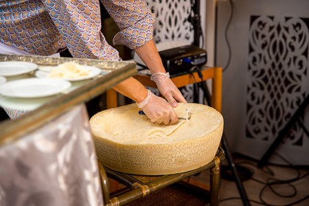 The cheese costs on a table. The person in gloves cuts out parmesan slices from it
