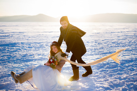 the groom rides the bride on the sledge on the stiffened lake against mountains. The bride in hands has a bouquet of flowersの写真素材