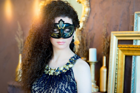 woman with black curly hair and a masquerade mask posing on a brown backgroundの写真素材