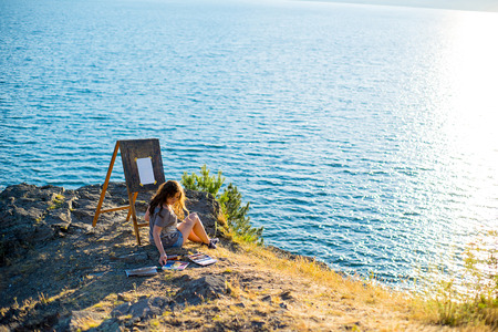 young artist draws a seascape at sunset on the highrockの写真素材