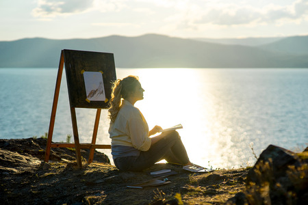 young artist draws a seascape at sunset on the highrockの写真素材