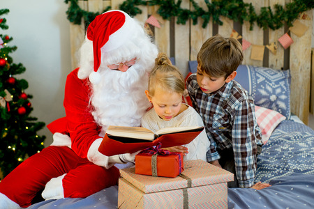 Santa Claus with two children reading a book near Christmas tree sitting on a decorated bedの写真素材