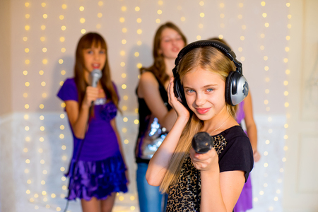 Group of happy girls singing together on karaoke on background of lights with first plan of one girlの写真素材