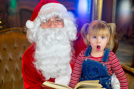 Santa Claus and girl sitting in Christmas room and reading a book. Christmas home decorの写真素材
