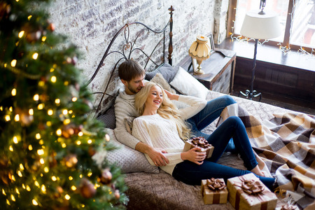 Happy couple of lovers in white sweaters give each other gifts lying on the bed. Christmas tree and new year gift at home. Young family togetherの写真素材