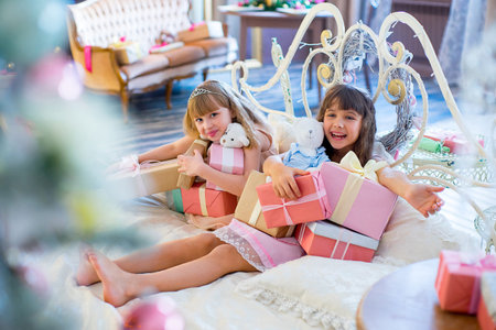 Two adorable girls in a white dress lying on the bed between Christmas giftsの写真素材