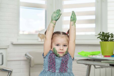 Little girl in mask paints with fingers.Little girl smiling, climbed up with her palms in green paint, self-isolation, coronavirus covid-19, stay homeの写真素材