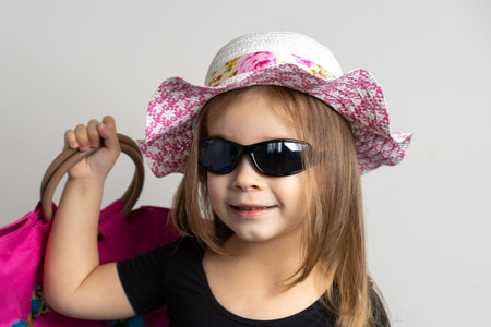 a cheerful little girl of 3-4 years old in sunglasses and a summer hat smiles happily and holds a...