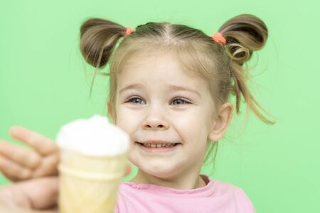 little girl 4 years old in a pink T-shirt on a green background holds out his hand to ice cream with delight, the child is happyの写真素材
