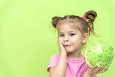 little girl 4 years old in a pink T-shirt on a green background holding a swing of cabbage in his hands and smiling. proper nutrition for preschoolers. vegetarian and kidsの写真素材