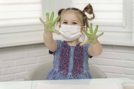 Little girl in mask paints with fingers.girl smiles and raised her green palms in the paint, self-isolation, coronavirus covid-19, stay homeの写真素材