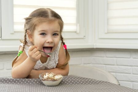 4 years old girl in a white blouse eagerly eats ice-cream from dumplings at a table in the kitchen and is happyの写真素材