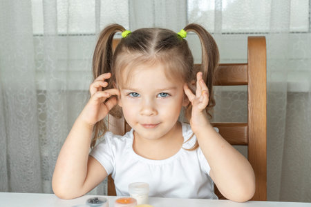3-4 years old girl with beautiful gray eyes ponders a drawing in front of paint cans. children's...