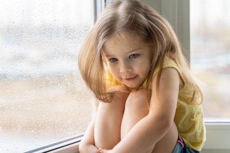 a little girl in a yellow blouse sits by the window with raindrops and is sad. children are bored at...