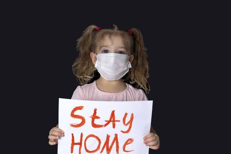 A little girl in a medical mask stands with a banner to stay home. A call to stay at home. holding a poster in front of him. quarantine. self-isolationの写真素材