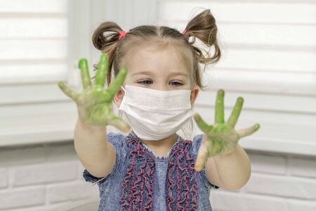 Little girl in mask paints with fingers.girl looks in surprise at her green palms in the paint, self-isolation, coronavirus covid-19, stay homeの写真素材