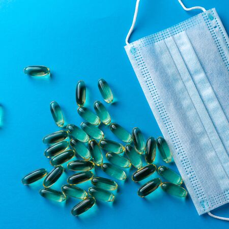 Surgical mask and a scattering of yellow fish oil capsules on a blue background. medical backgroundの写真素材