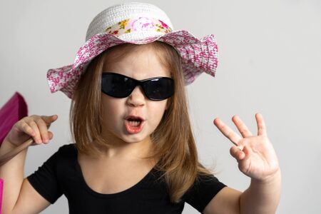 a cheerful little girl of 3-4 years old in sunglasses and a summer hat smiles happily and grimaces and holds a beach bag in her hand. summer holiday conceptの写真素材