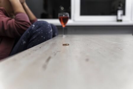 the girl is sitting at the table with her hands on her head, in front of her is a bottle and a glass of wine. there is a wedding ring on the table. Depression due to divorce.の写真素材
