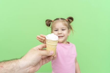 little girl 4 years old in a pink T-shirt on a green background holds out his hand to ice cream with delight, selective focus on ice cream, child in defocusの写真素材