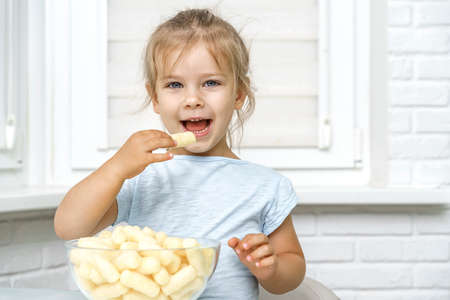 4 years old girl sits at the kitchen table and happily eats corn sticks from a glass cupの写真素材