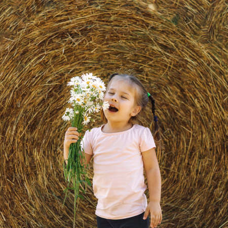 little girl 4 years old with a bouquet of field daisies sat down to rest near a haystack on a summer dayの写真素材