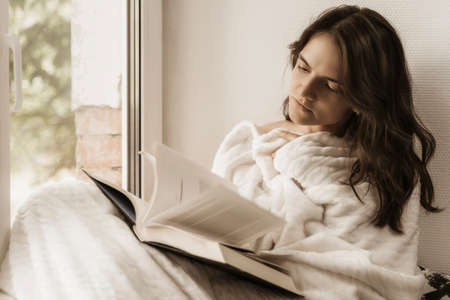 Relaxing and thinking woman. Calm woman with a book sits on the windowsill at home.の写真素材