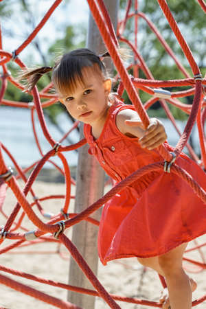 little girl in a red dress perch on a web rope-ladder structure in a children's playground for fun climbing.の写真素材