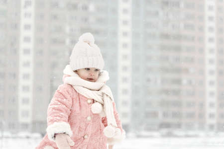 beautiful little girl in a pink fur coat, hat and scarf, against the background of multi-storey buildings in a snowfallの写真素材