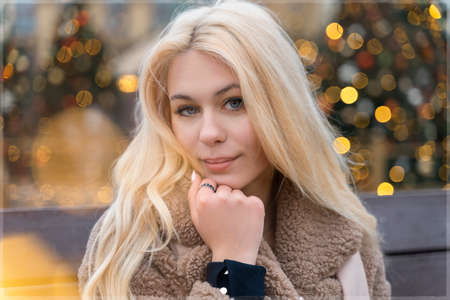 01 14 2020 Moscow, Russian Federation. beautiful young blonde woman in a fur coat made of faux fur sits on a bench near a Christmas tree smiling. new year and christmas conceptのeditorial素材