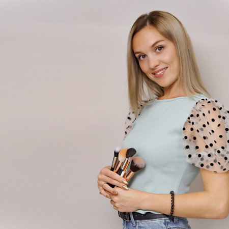 Makeup concept. Smiling blond woman holding cosmetic brushes, posing on a light background. Studio...