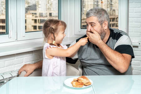 a cute little girl laughingly presents cooked pancakes to her gray-haired dad. father jokingly refuses to eat. family values and the concept of joint leisureの写真素材