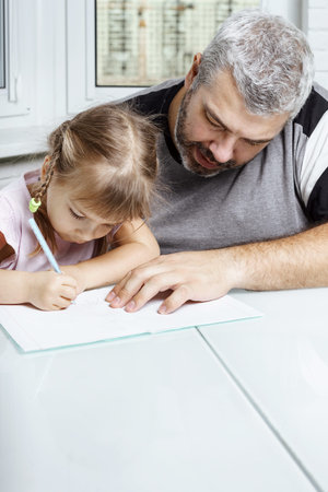 adult gray-haired father teaches little daughter to write at the table at home. family values...