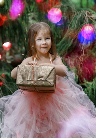 A cute little girl in a pink dress stands by the Christmas tree and tries to guess what is in the...