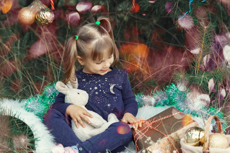 A cute little girl in a pink dress stands by the Christmas tree and tries to guess what is in the...
