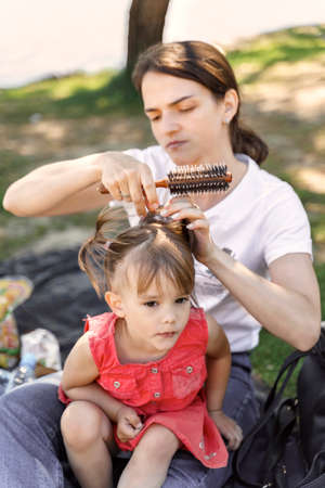young Mom Brushing Little Daughter Outdoor. young woman doing her little daughter's hairstyleの写真素材