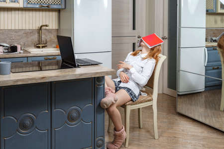 girl student on an online lesson in the kitchen. the child leaned back in the chair and covered her face with a notebook. children are tired of distance learning. e-learning in connection with the coronavirus.の写真素材