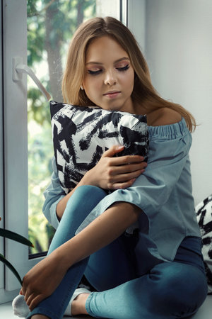 beautiful sad girl sitting by the window and hugging a pillow.