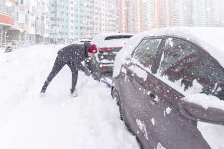 a young man in a warm jacket and hat is digging his car out of the snow captivity with a shovel. heavy snowfall, poor visibilityの写真素材